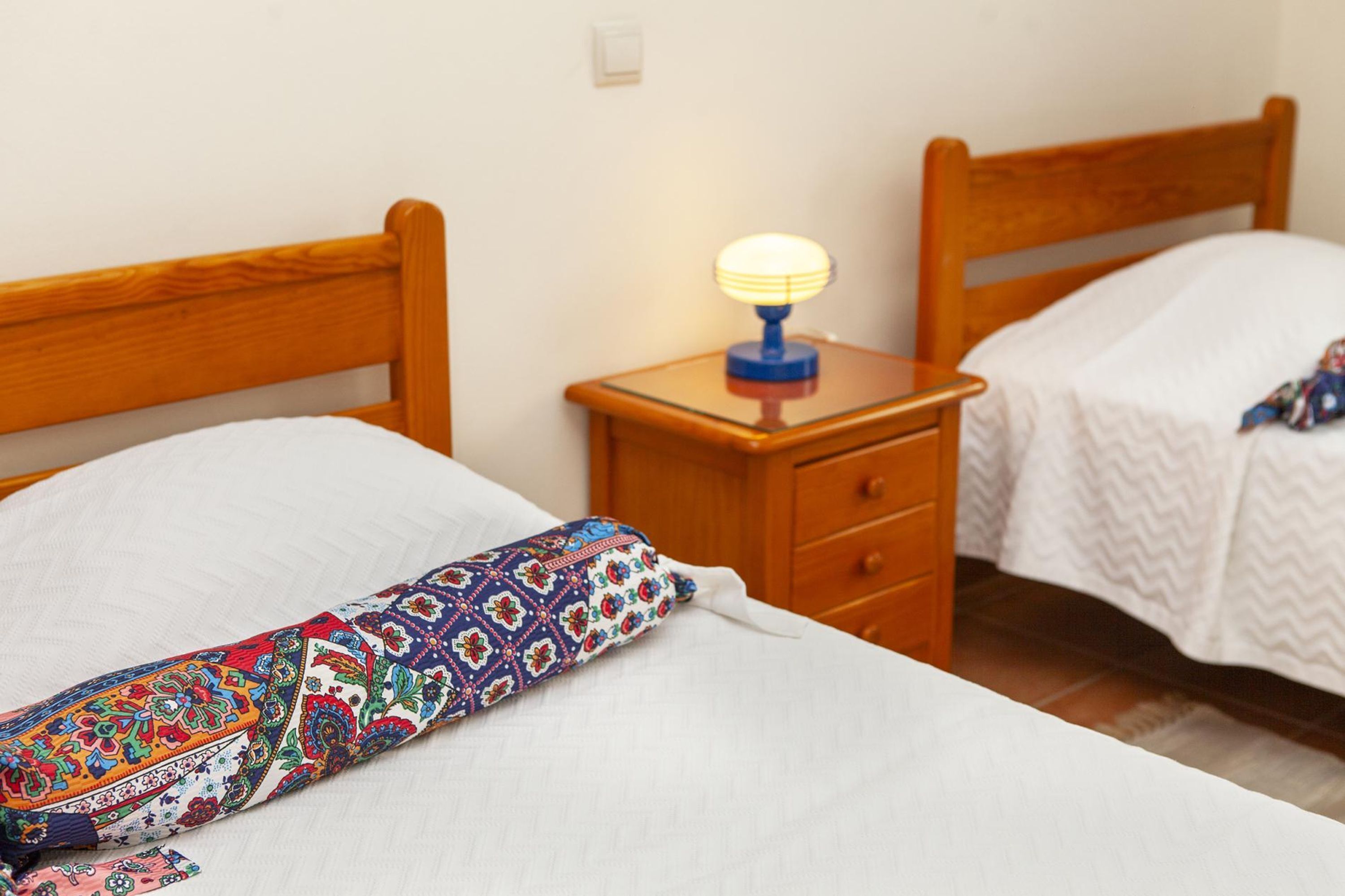 Un dormitorio con dos camas de madera, una mesita de noche con lámpara y un cojín colorido.