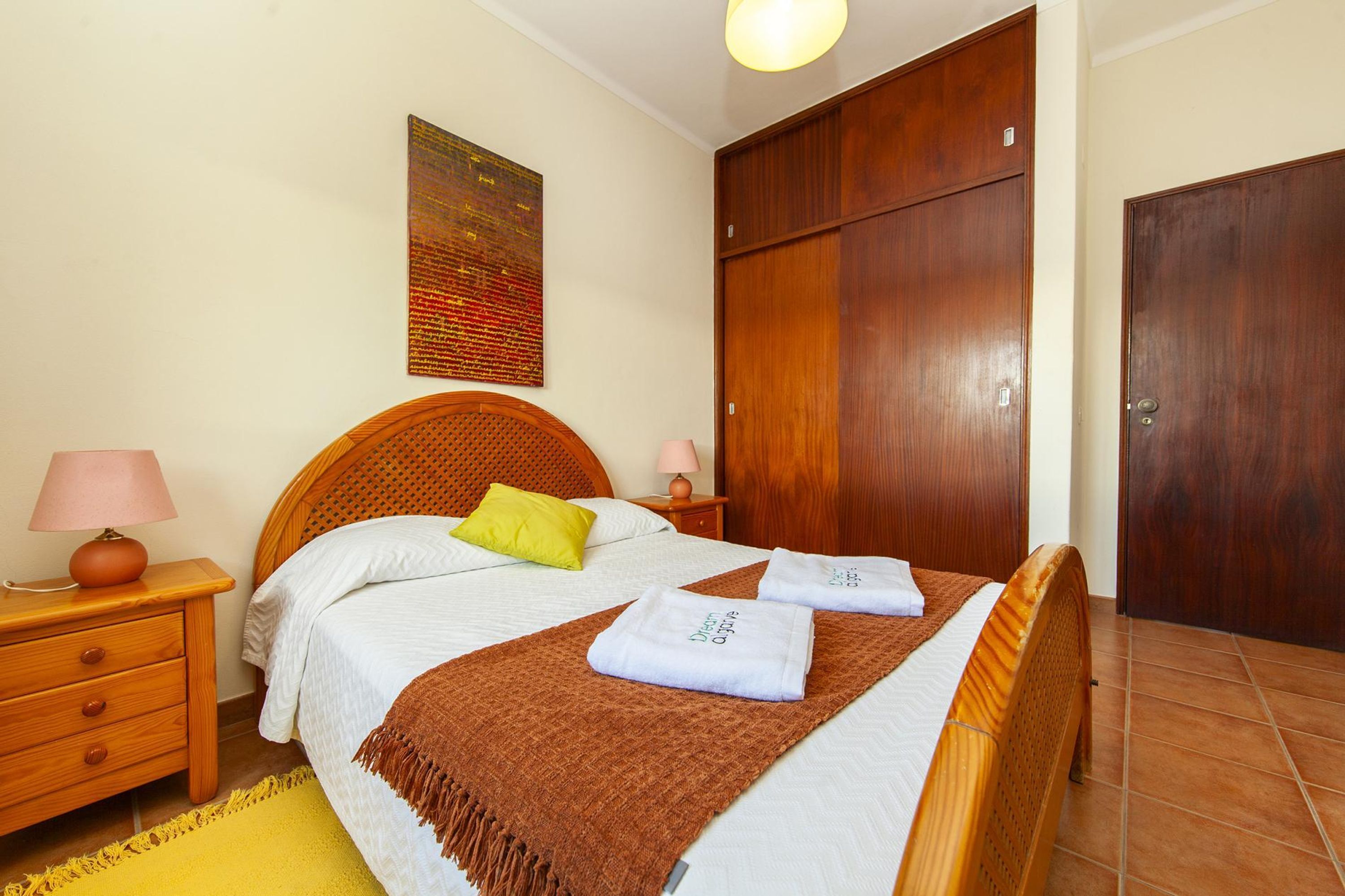 Un lit bien fait avec une tête de lit marron, une table de chevet en bois avec une lampe, un tapis jaune et une armoire en bois.
