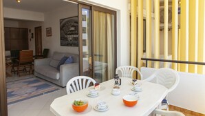 A dining area with a table set for two, a sofa, and a sliding glass door leading to a balcony with yellow vertical blinds. - A02 - Casa Topazio in Quarteira (Loulé)