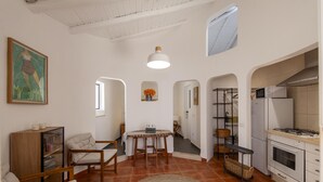 Een ruime woonkamer met een eettafel, stoelen, een keuken met witte apparatuur en een plafond met zichtbare balken.