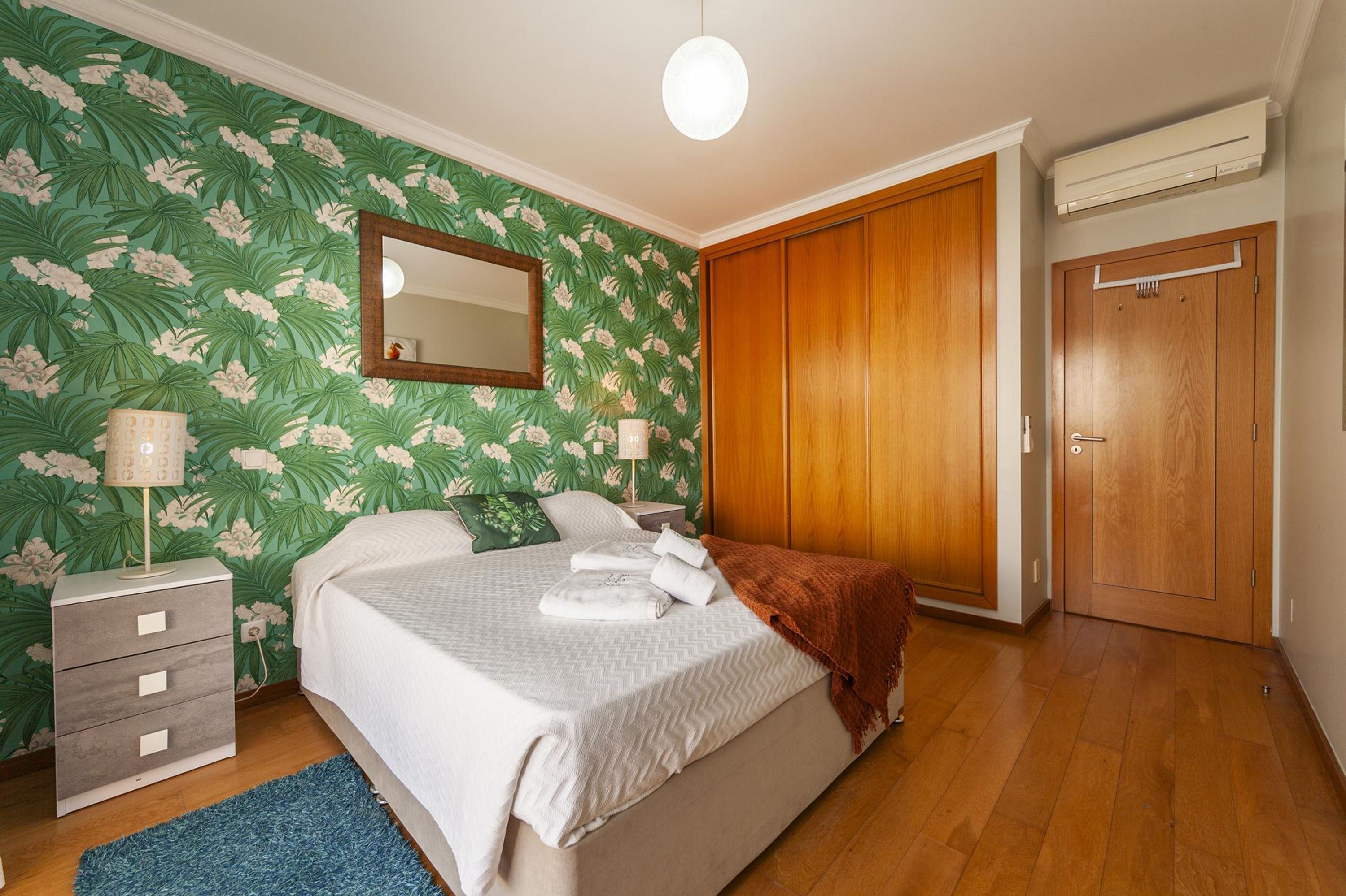 A bedroom with a bed, a nightstand, a mirror, a wardrobe, and a wall with floral wallpaper.