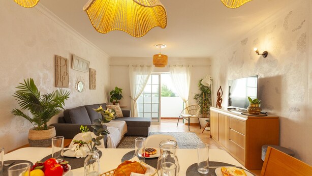 A modern living room with a sofa, dining table set with food, and a kitchen area with cabinets and a sink. - B01 - Mar a Vista Rocha Apartment (Portimão)