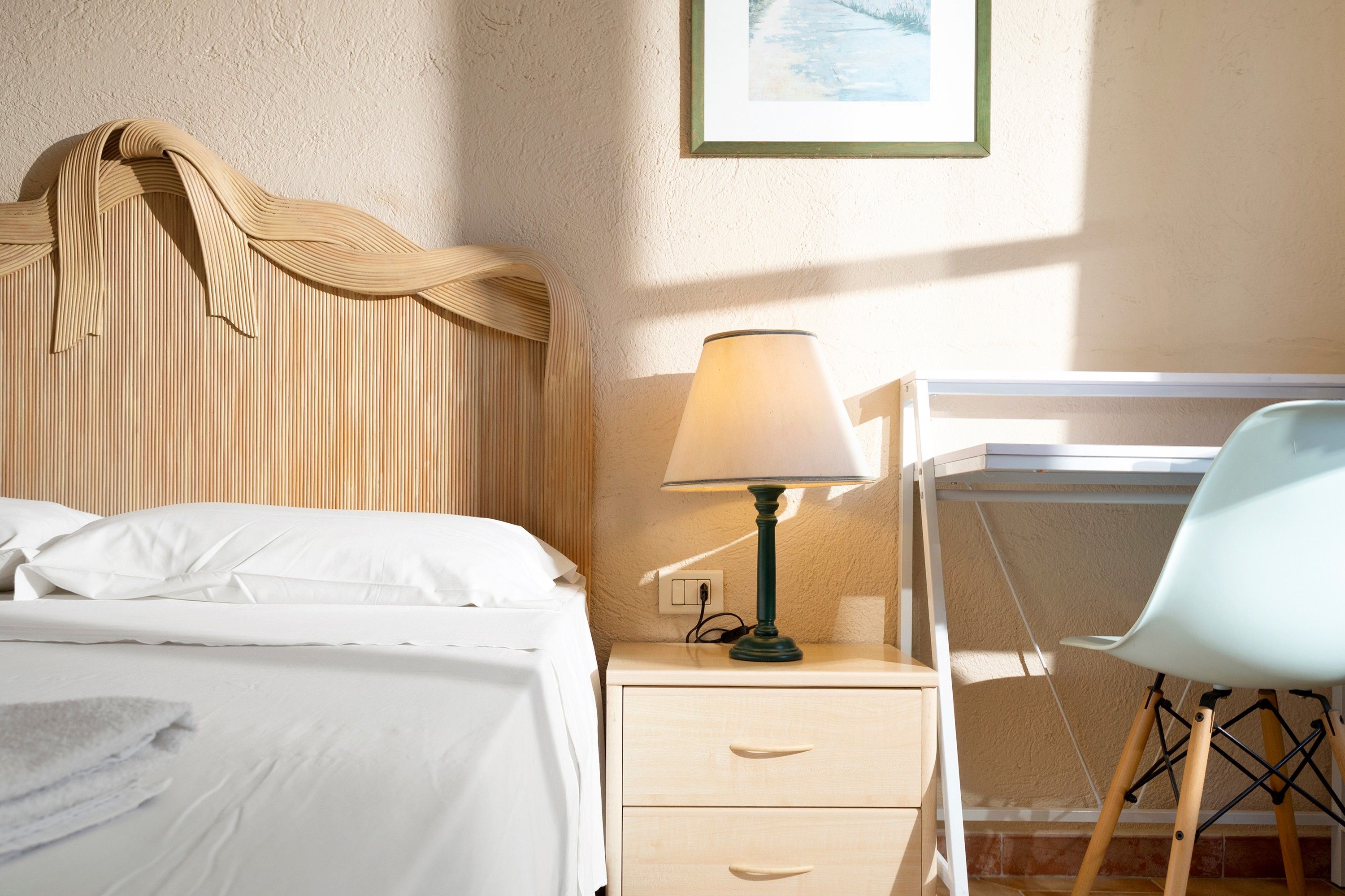 A neatly made bed with white linens, a bedside lamp, a wooden nightstand with drawers, and a chair with a light blue cushion.