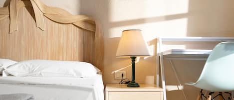 Una cama bien hecha con sábanas blancas, una lámpara de noche, una mesita de madera con cajones y una silla con cojín azul claro.