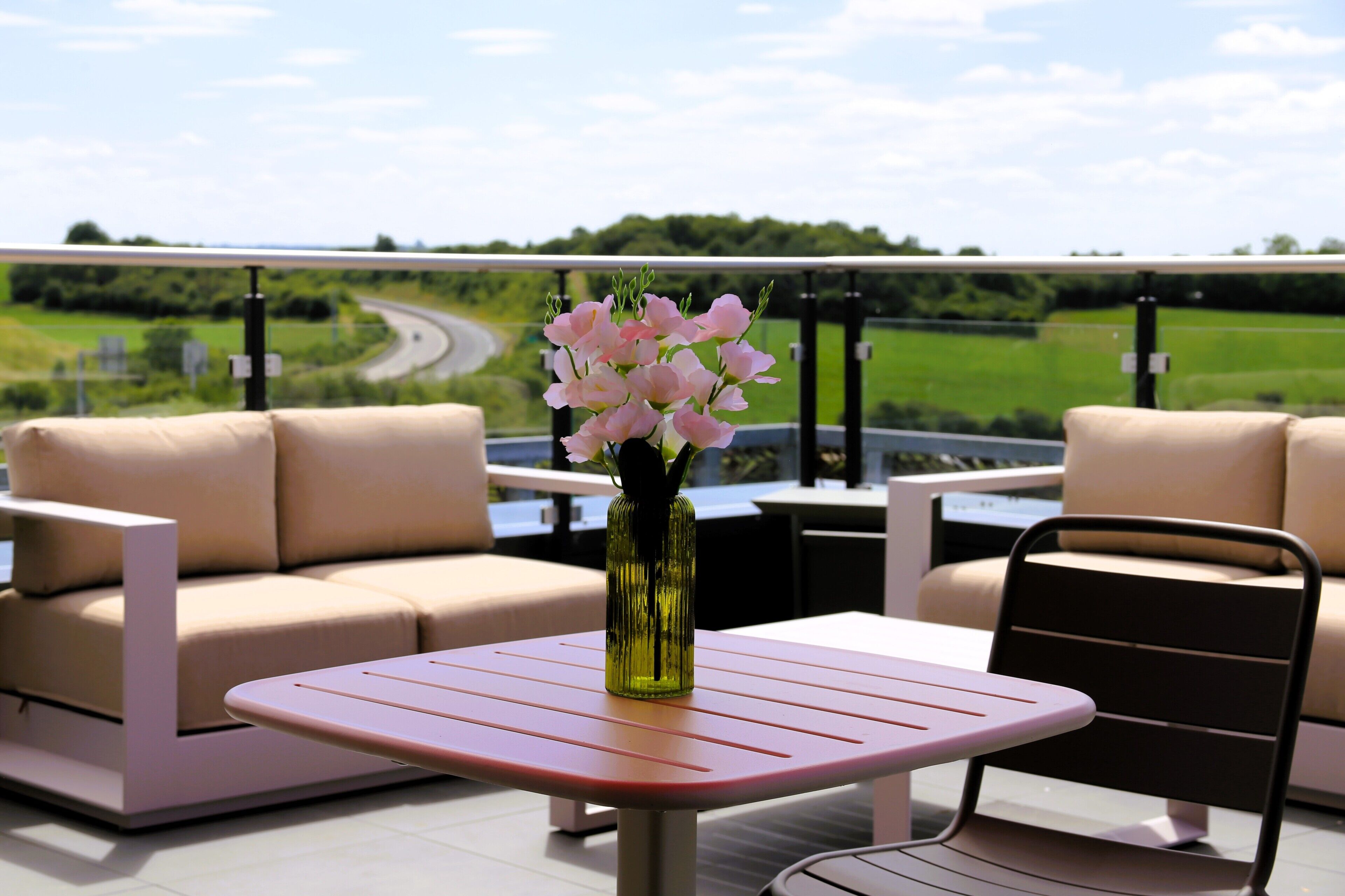 A patio area with a table and a sofa, a vase with flowers, and a view of a road and greenery.