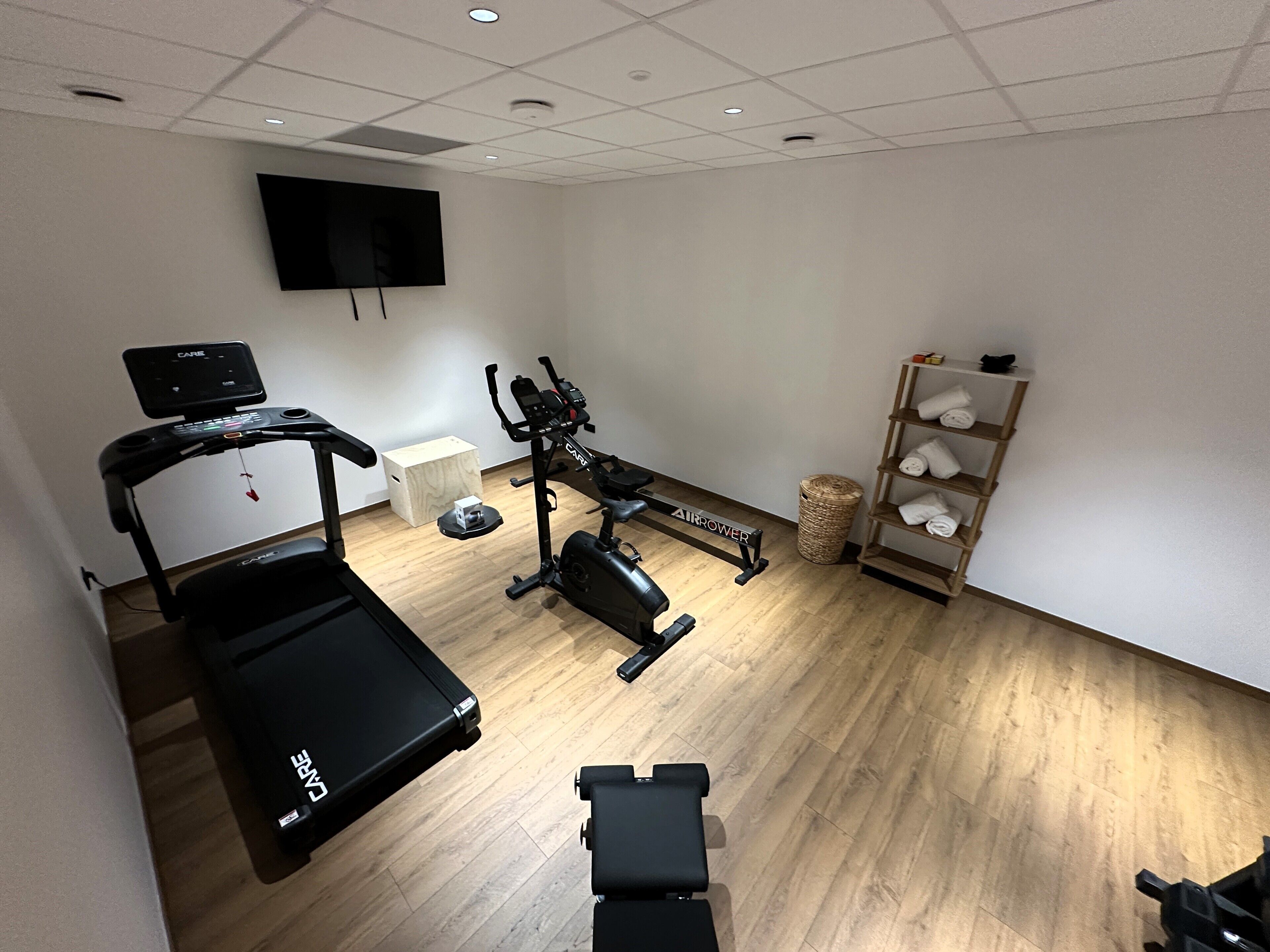 A gym with cardio equipment, a wall-mounted TV, and a shelf with towels.