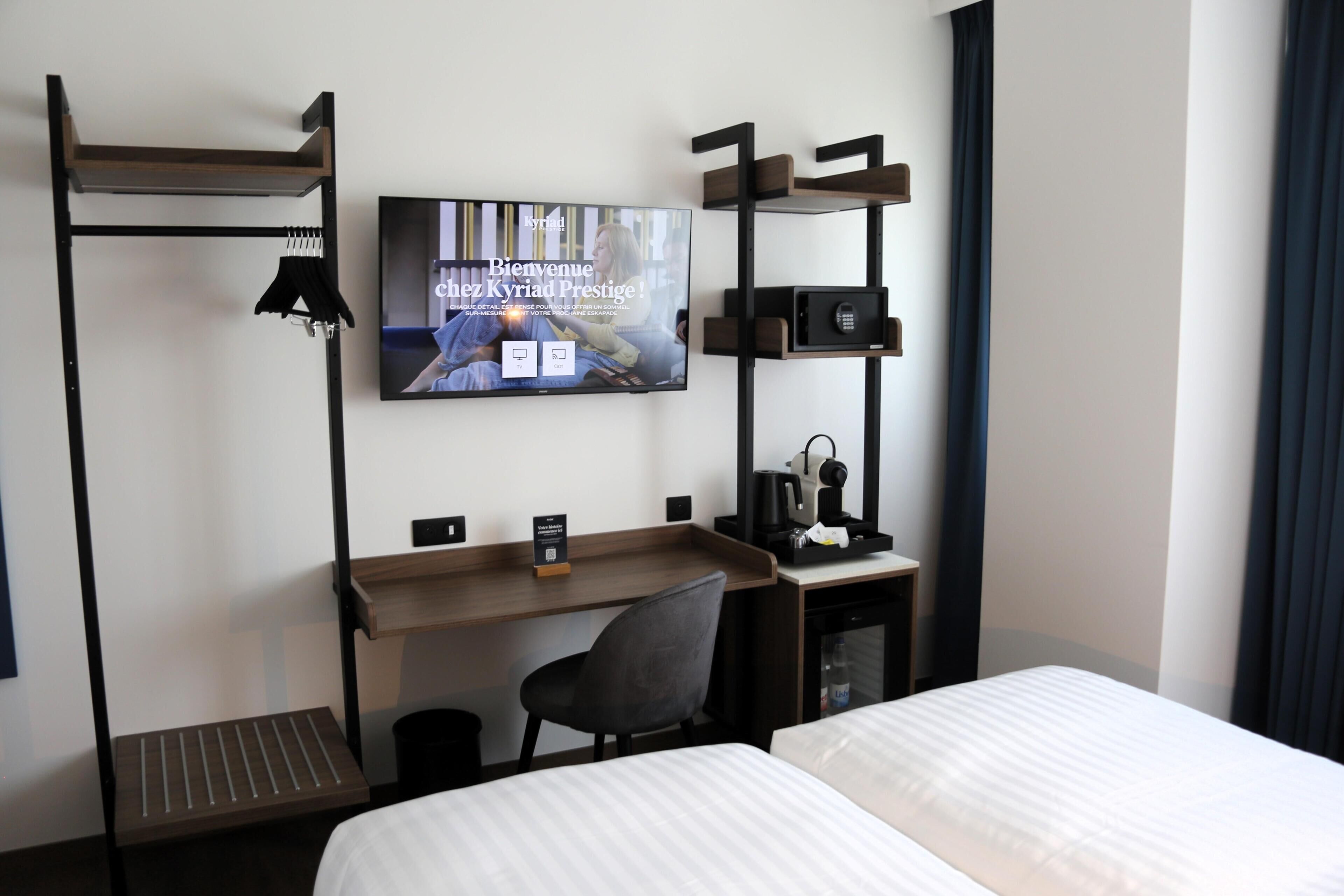 Una habitación de hotel con cama, escritorio con silla, televisor montado en la pared y un armario pequeño.