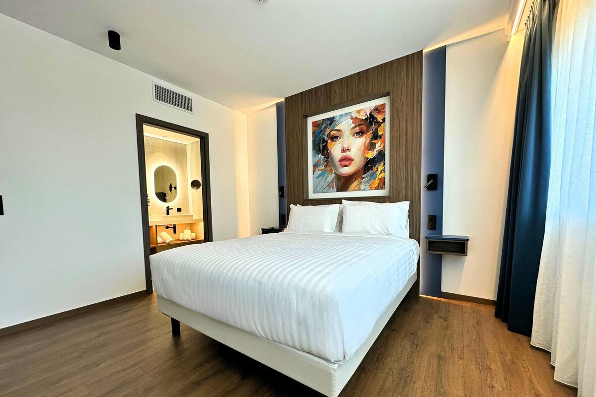 A hotel room with a large bed, a bathroom with a mirror and sink, and a painting on the wall.