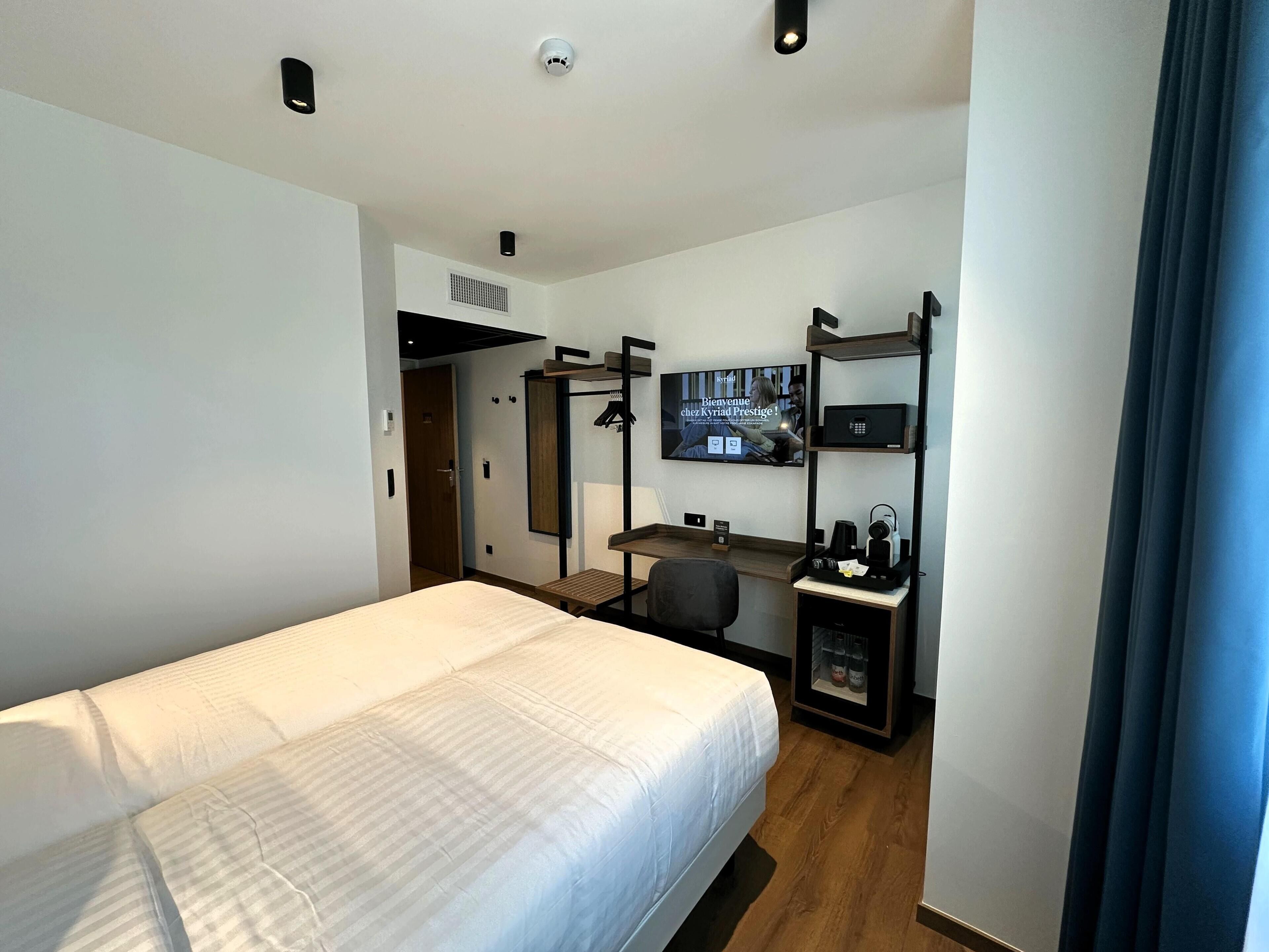 A modern hotel room with a bed, desk, chair, TV, and a small shelf with amenities.
