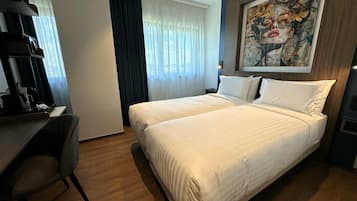 Quarto de hotel com cama, escrivaninha, cadeira, um quadro na parede e janela com cortinas.