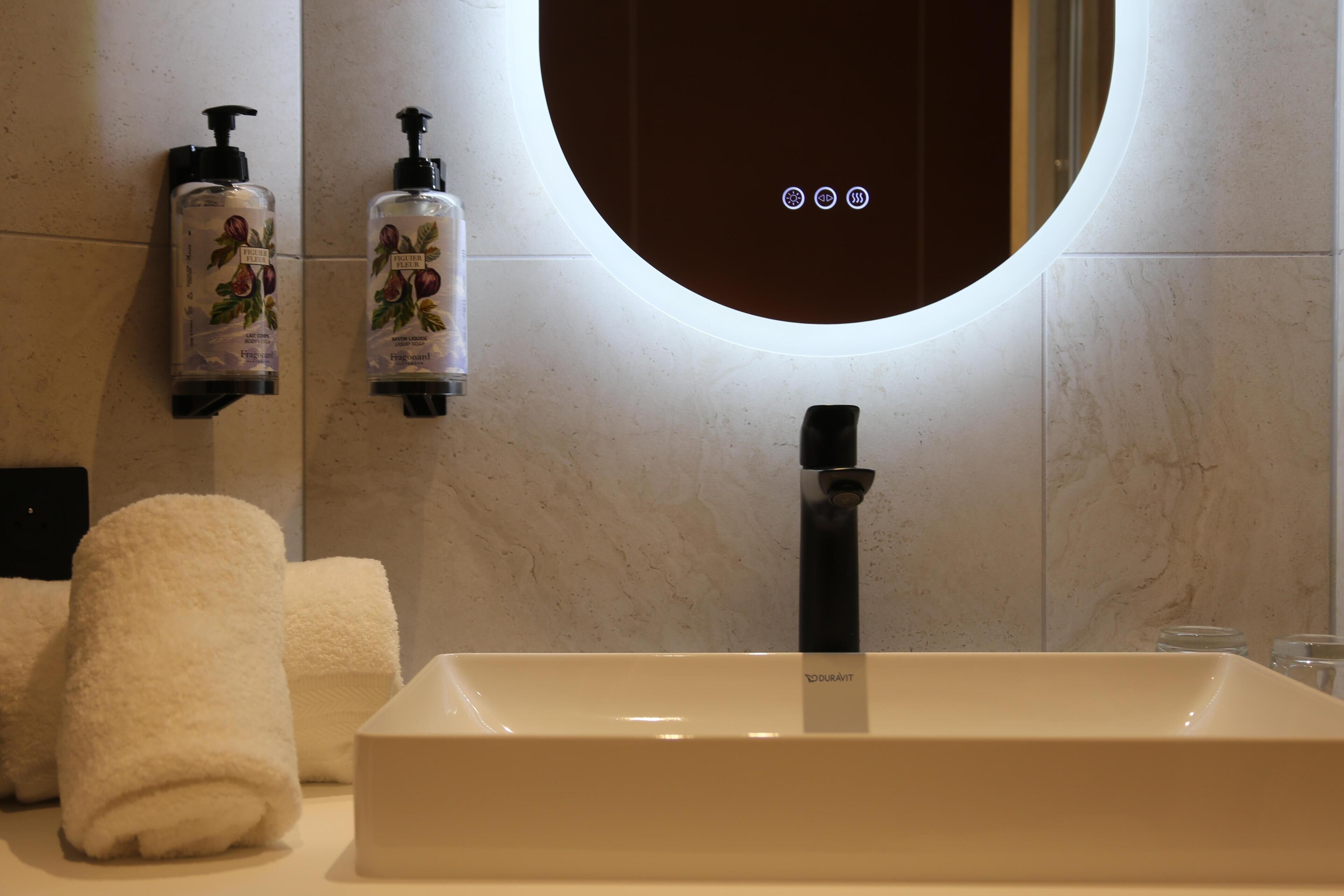 Un baño con un lavamanos blanco, dos dispensadores de jabón, un espejo y una toalla.