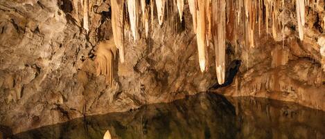 Interior de cueva con estalactitas y estalagmitas, un charco de agua reflectante y suelo rocoso.