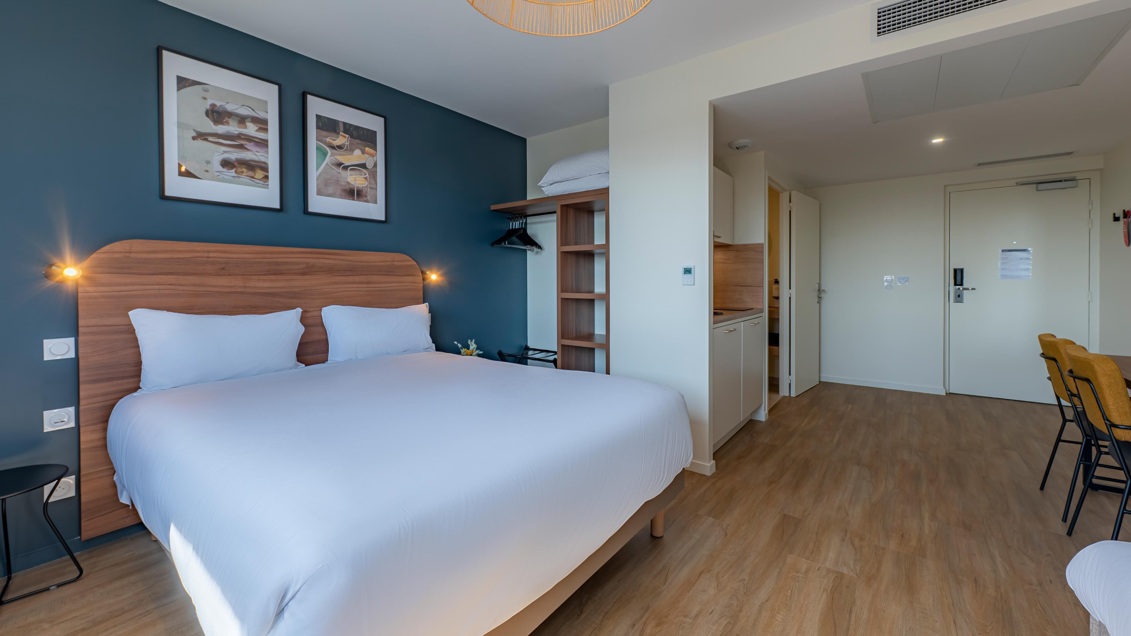 A hotel room with a large bed, a desk, a chair, a wardrobe, and a kitchenette.