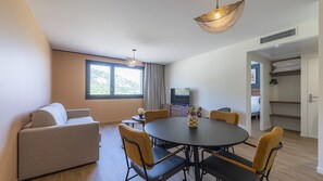 A modern living room with a sofa, dining table, chairs, a TV, and a large window with a mountain view.