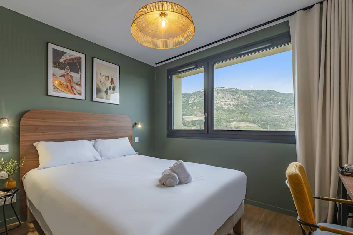 A hotel room with a large bed, a wooden headboard, a window with a view of hills, and a table with a chair.