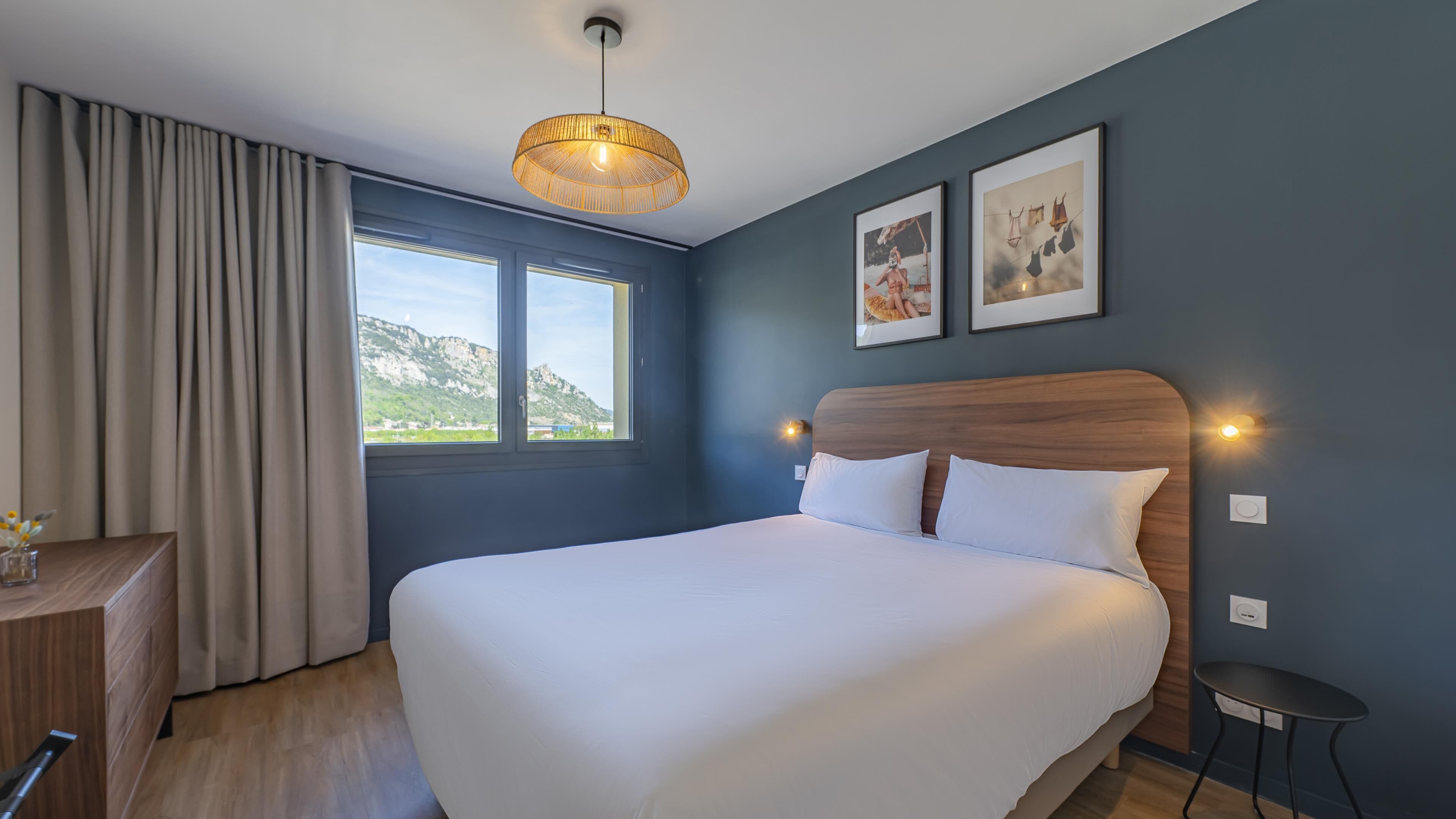 A hotel room with a large bed, a desk, a chair, a window with a view of mountains, and two framed pictures on the wall.