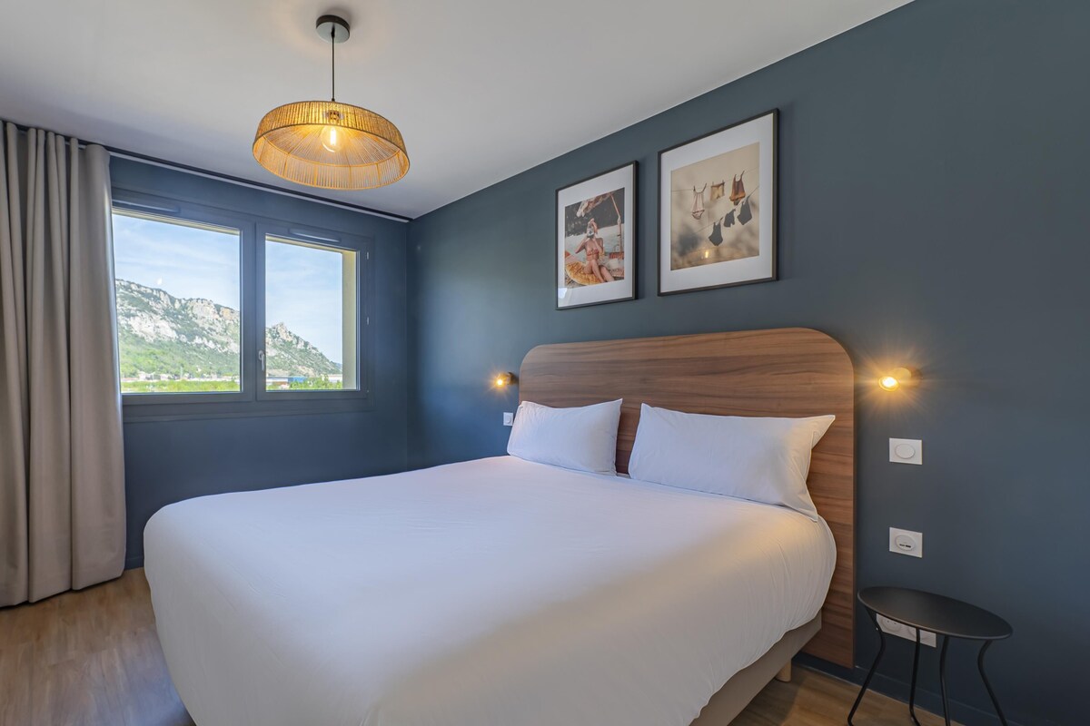 Ein Hotelzimmer mit einem großen Bett, einem Schreibtisch, einem Stuhl, einem Fenster mit Bergblick und zwei gerahmten Bildern an der Wand.