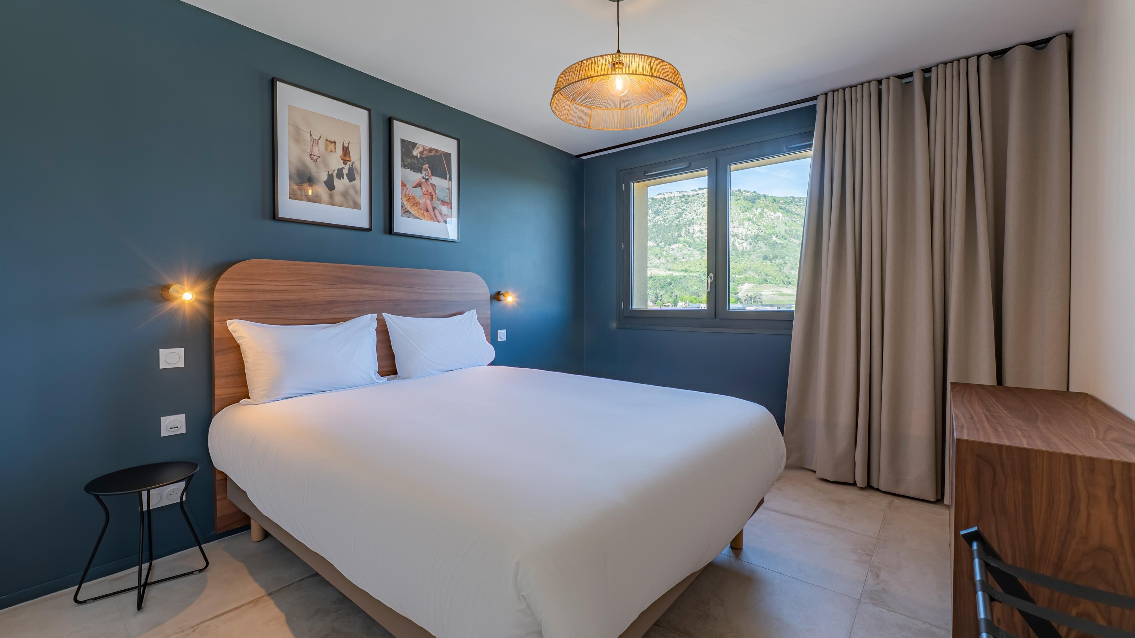 A bedroom with a bed, a bedside table, a lamp, a window with curtains, and two framed pictures on the wall.
