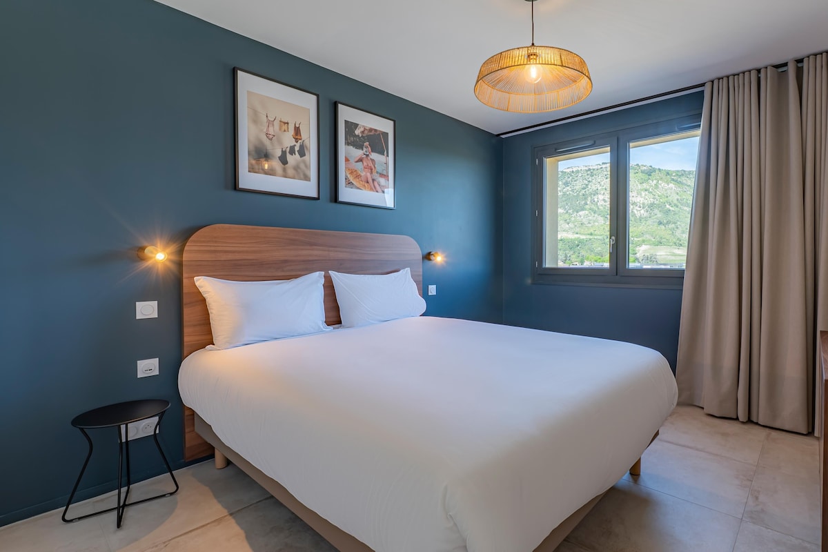 Ein Schlafzimmer mit einem Bett, einem Nachttisch, einer Lampe, einem Fenster mit Vorhängen und zwei gerahmten Bildern an der Wand.