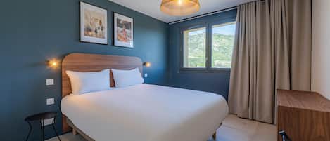 A bedroom with a bed, a bedside table, a lamp, a window with curtains, and two framed pictures on the wall.