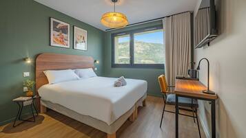 Habitación de hotel ordenada con cama, escritorio, silla, televisión y ventana con vistas al exterior.