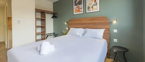 Una cama bien hecha con ropa blanca y una toalla doblada, cabecera de madera, mesita de noche pequeña y dos cuadros en la pared.