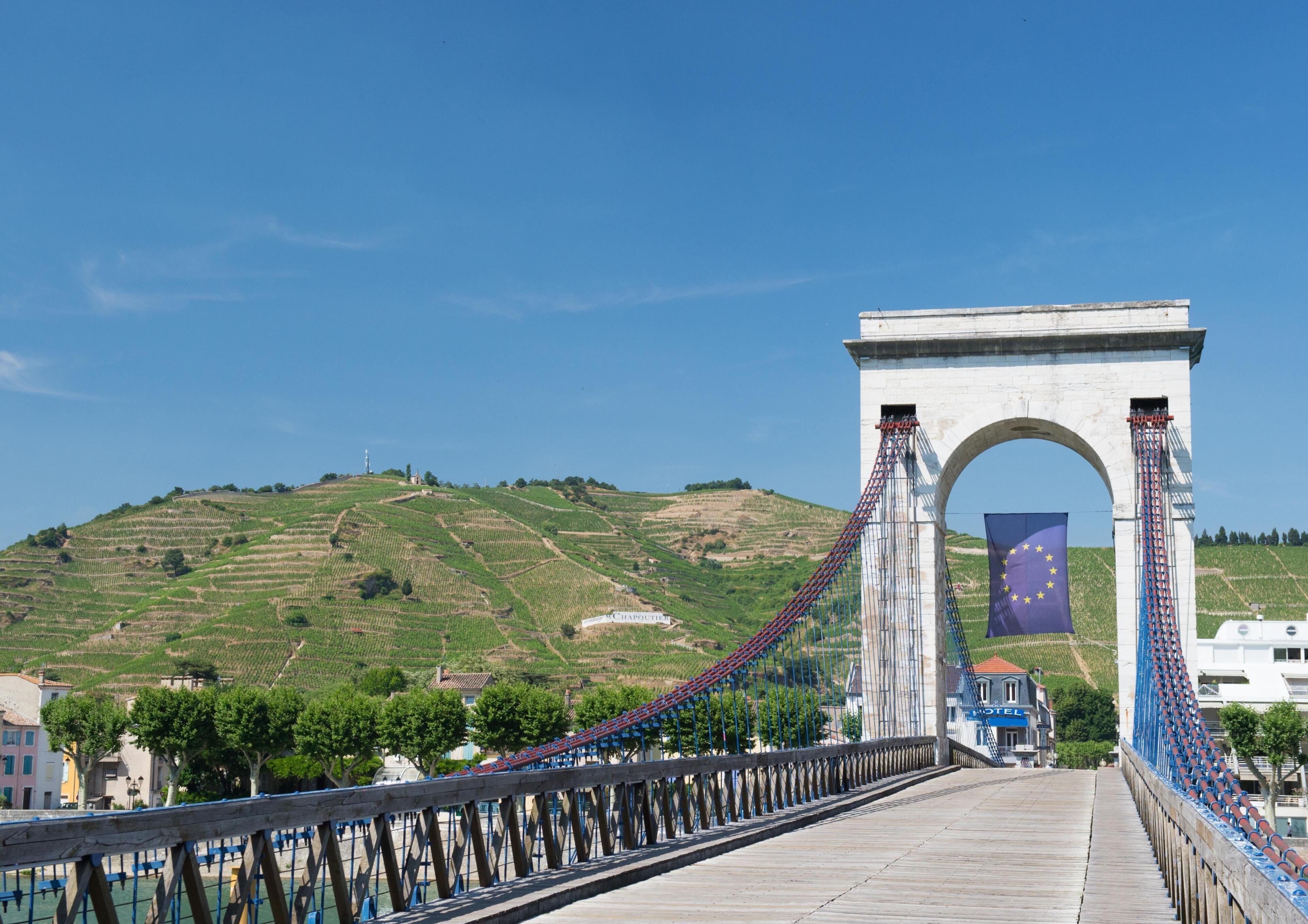 A bridge with an arch and a flag, vineyards in the background, and a hotel with a sign.