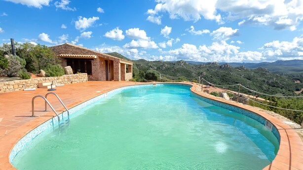 Ein Pool mit einer Steindeck, ein kleines Gebäude mit einem Ziegeldach und eine gebirgige Landschaft im Hintergrund.