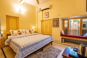 A bedroom with a large bed, a window with a view, a bedside table with a kettle, and a small table with a lamp. - HOTEL HIMMAT NIWAS (Jaisalmer)