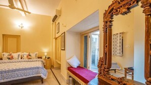 A bedroom with a large bed, a small bench, a wooden chair, a large mirror, and a window with curtains. - HOTEL HIMMAT NIWAS (Jaisalmer)