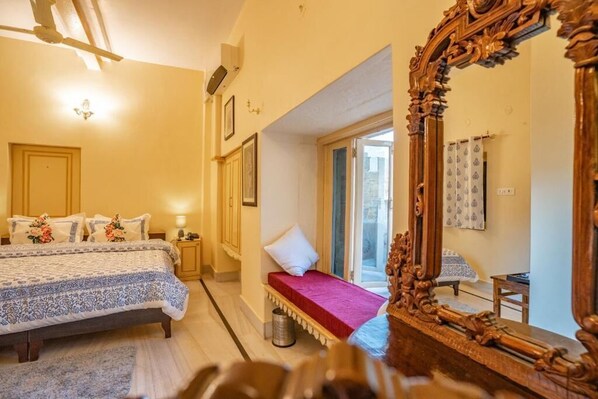 A bedroom with a large bed, a small bench, a wooden chair, a large mirror, and a window with curtains. - HOTEL HIMMAT NIWAS (Jaisalmer)