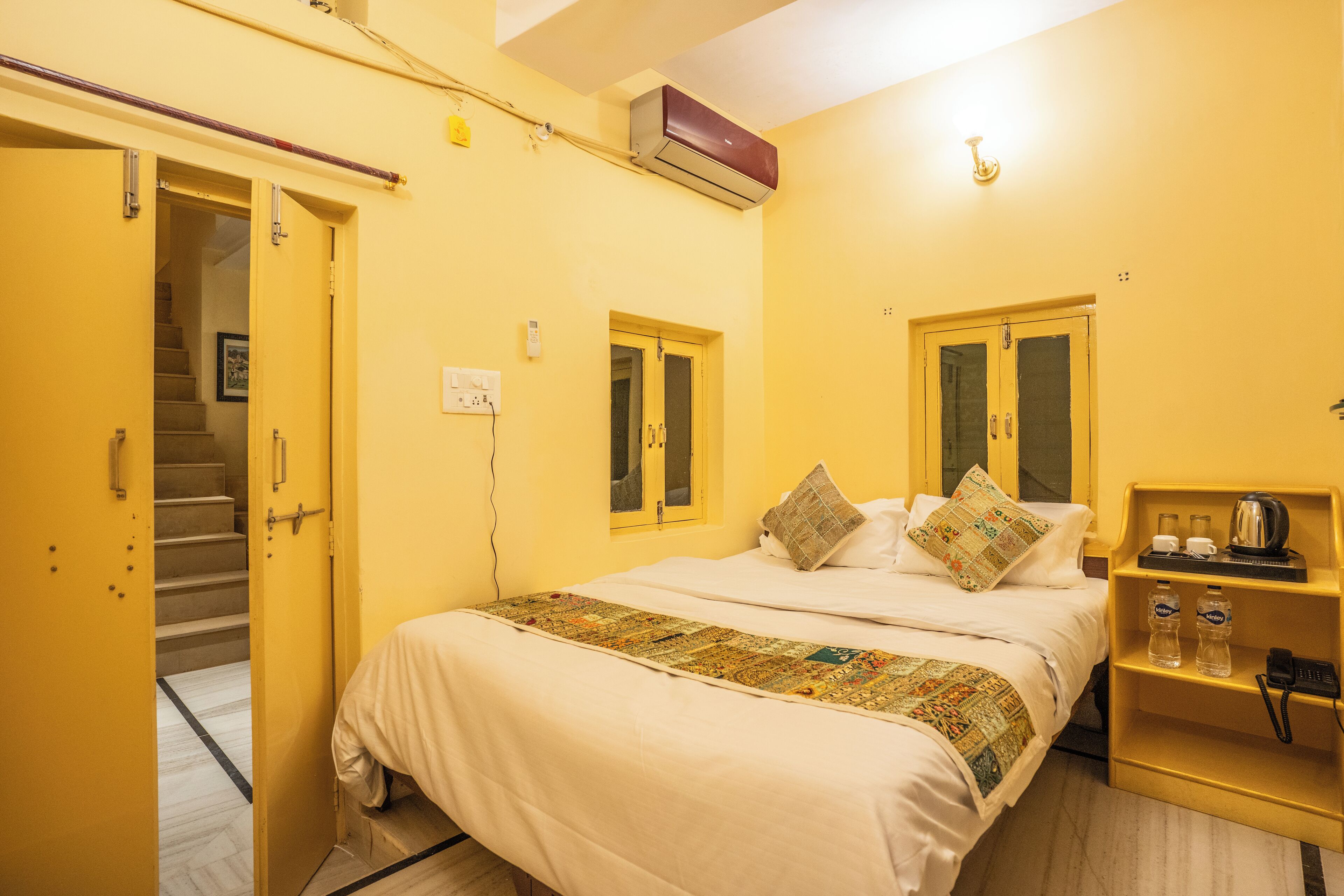 A hotel room with a bed, a bedside table with a kettle, and a shelf with water bottles.