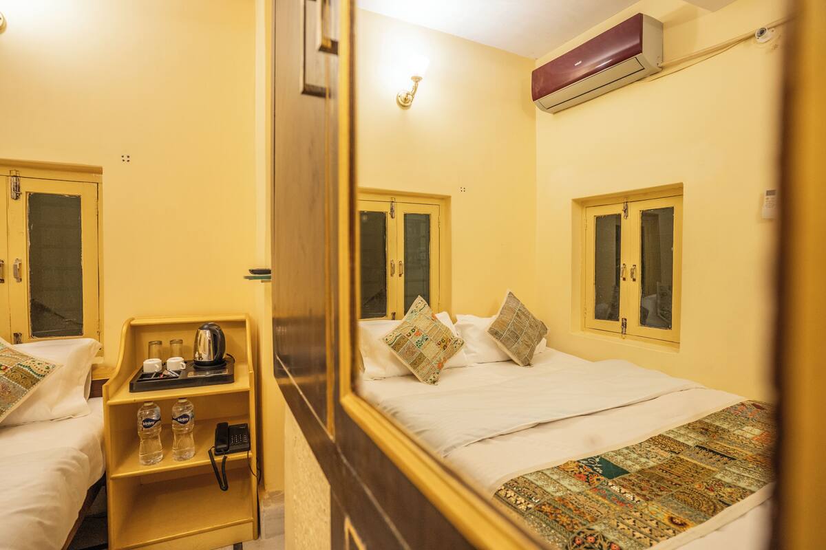 Habitación de hotel con una cama, una mesita de noche con tetera, tazas y botellas, y un espejo que refleja la habitación.