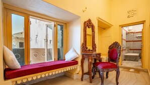 A room with a red cushioned bench, a wooden chair, a mirror, and a bathroom visible through a glass door. - HOTEL HIMMAT NIWAS (Jaisalmer)