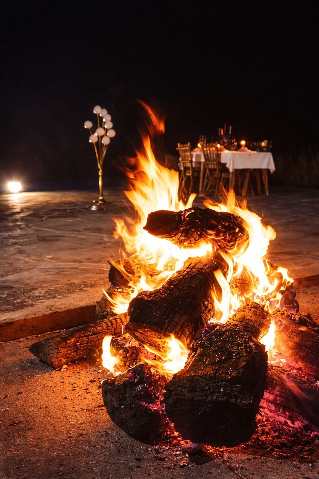 A bonfire with logs and flames, a dining table with chairs, and a white tablecloth in the background.