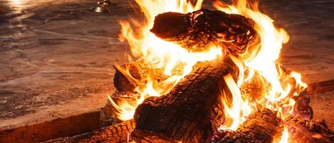 A bonfire with logs and flames, a dining table with chairs, and a white tablecloth in the background.