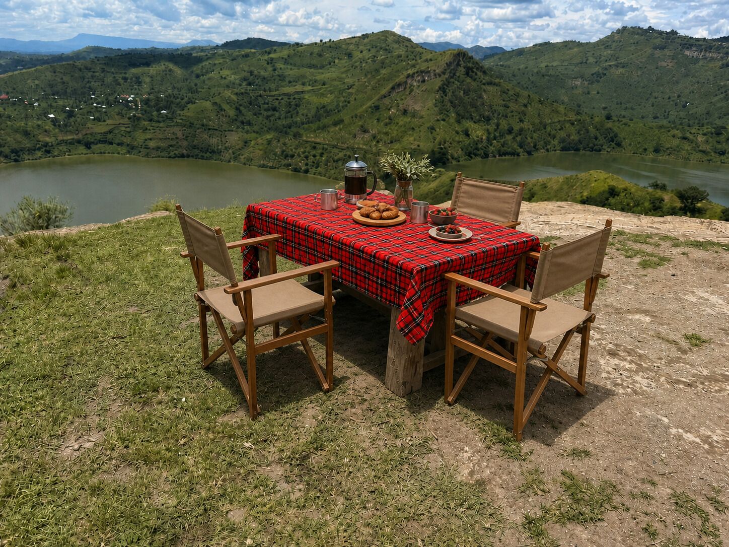 A table set on a grassy hill with a red plaid tablecloth, surrounded by wooden chairs, overlooking a lake and mountains.