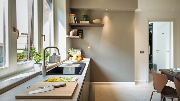 Une cuisine moderne équipée d’une cuisinière intégrée, d’un évier et d’une planche à découper en bois. Des étagères au-dessus du plan de travail servent à ranger des livres et des objets décoratifs.