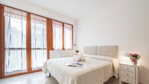 Un dormitorio con una cama grande, una mesita de noche con una lámpara y un jarrón de flores, y una ventana con cortinas traslúcidas.
