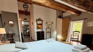 A large bed with white linens and a textured pillow, a wooden ceiling, a mirror, and a door leading to another room. - Mas de la Meï (Cavaillon)