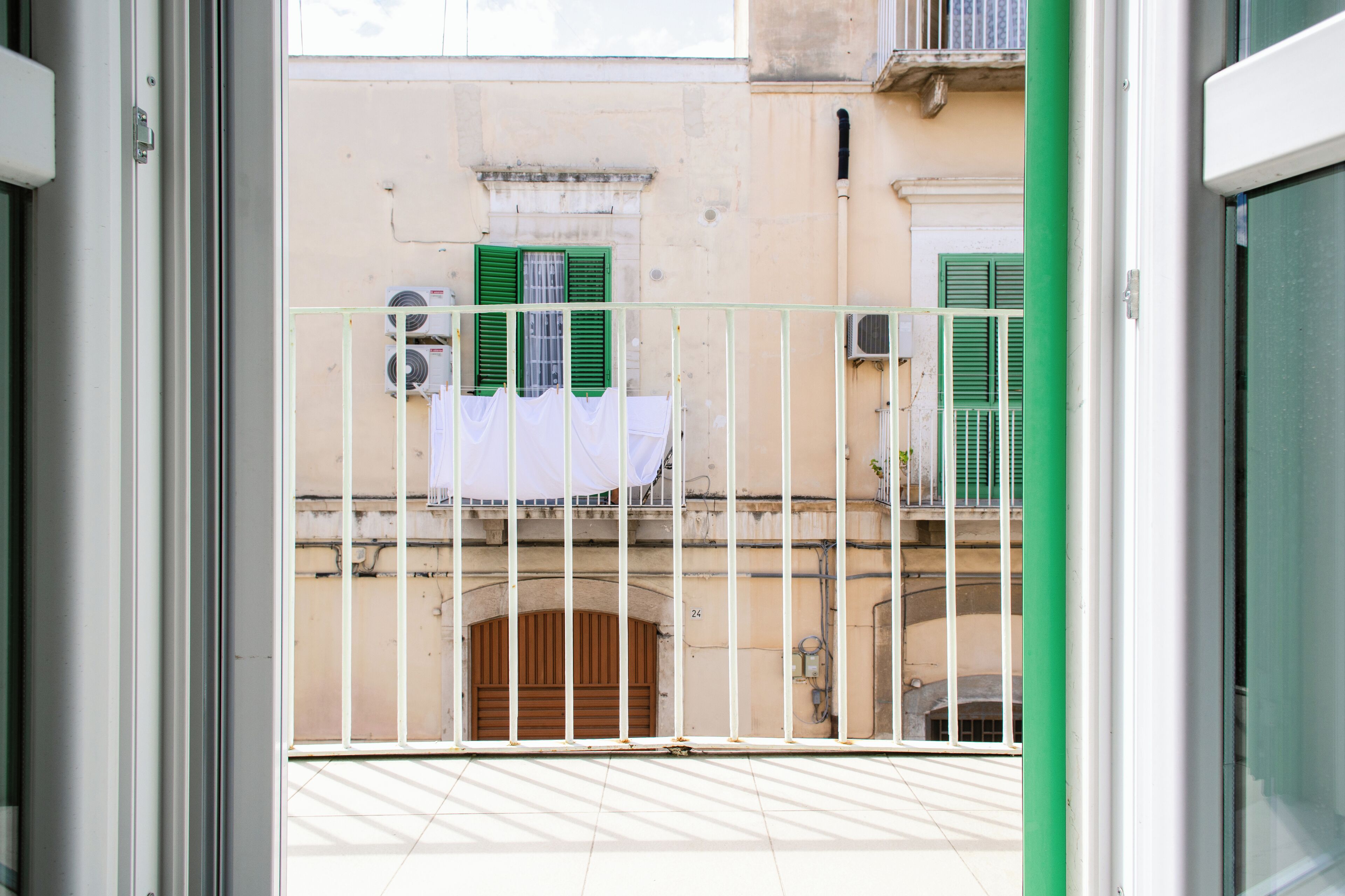 A balcony with a metal railing, a view of a building with green shutters, and a white cloth hanging on the railing.