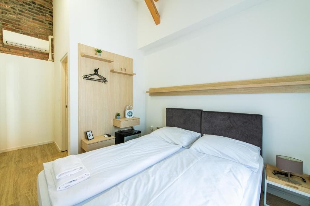 A neatly made bed with white linens, a wooden headboard, a bedside table with a lamp, and a small shelf with a plant and a clock.