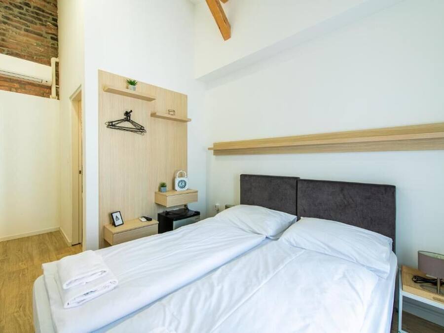 A neatly made bed with white linens, a wooden headboard, a bedside table with a lamp, and a small shelf with a plant and a clock.