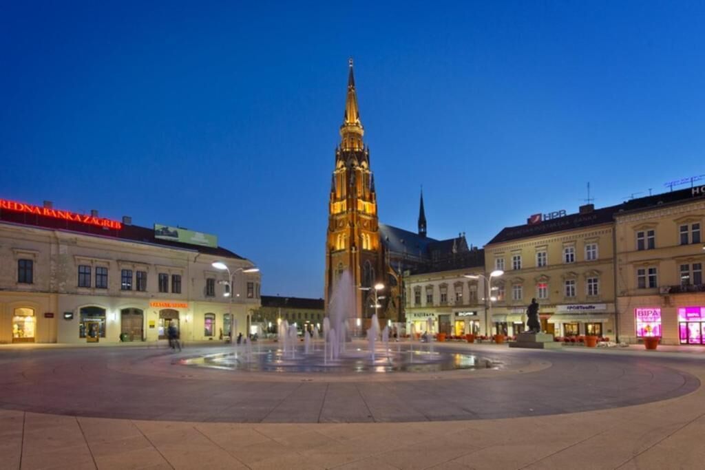 A square with a central fountain, surrounded by historic buildings with illuminated signs.