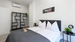 A modern bedroom with a large bed, a black metal shelving unit, a small round table with a clock, and a wall-mounted picture. - City Rooms OS (Osijek)