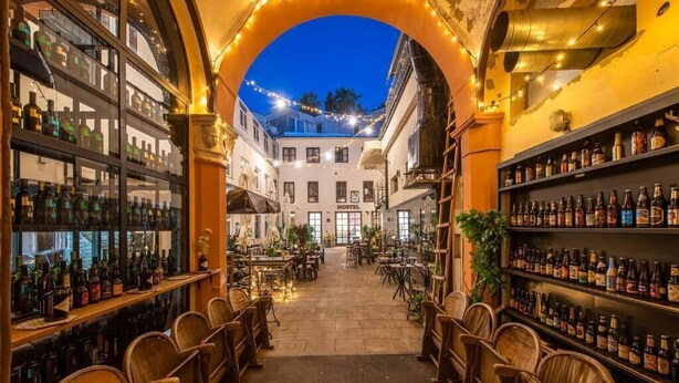 An indoor bar area with wooden chairs and shelves of bottles, an arched entrance, and a view of the street outside. - City Rooms OS (Osijek)