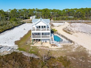Een twee verdiepingen tellend strandhuis met zwembad, omgeven door zand en begroeiing.