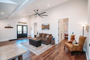 A spacious living room with a large ceiling fan, a brown sofa, a wooden coffee table, and a patterned rug. - Owls Nest: Downtown stay, Deck, Hot Tub, and Wifi (Buena Vista)
