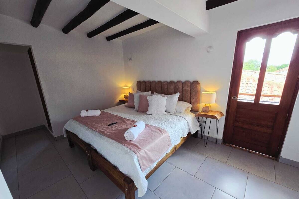 Une chambre à coucher avec un lit en bois, deux tables de chevet avec des lampes, une grande fenêtre et une porte donnant sur l’extérieur.