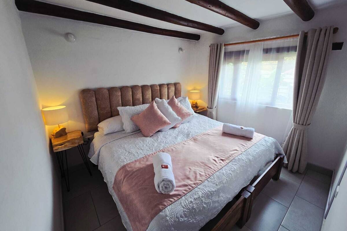 Un lit bien fait, avec des oreillers et une serviette roulée, une tête de lit en bois, des lampes de chevet et une fenêtre avec des rideaux.